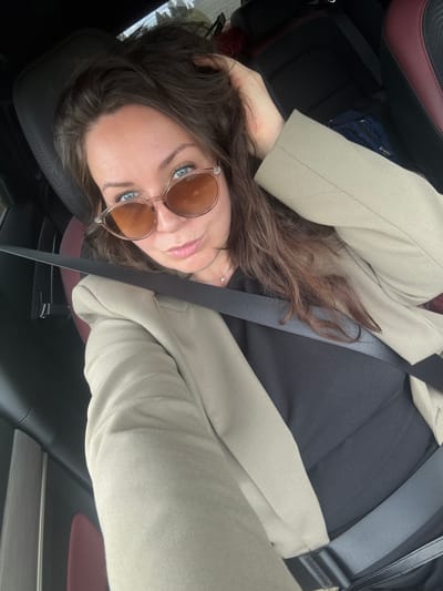 The image shows a woman in the driver's seat of a car. She is wearing sunglasses and a light-colored blazer over a black top. Her left hand is resting on her head. The car's interior is visible, with a dark dashboard and seats, and a burgundy accent on the passenger seat. A seatbelt is fastened across her chest. The background outside the car appears to be blurred foliage. There is no indication of the location being in Krasnoyarsk, Russia.