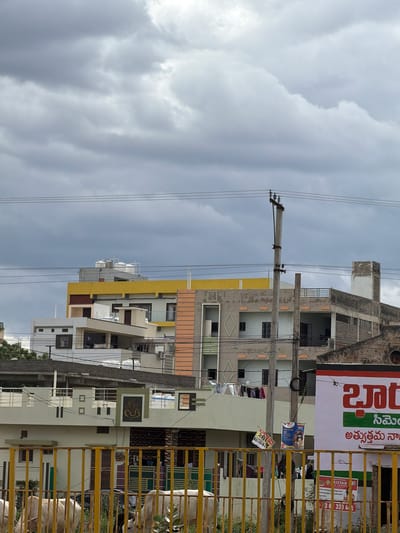 The image captures a cloudy sky over a cityscape, likely in B Camp, India. In the foreground, a yellow metal fence separates the viewer from a scene with two light-colored cows grazing, one with its head down. Behind the fence, several multi-story buildings are visible. One building has a prominent yellow band across its top. Another building features a concrete utility pole with electrical wires running across the frame, along with several pieces of laundry hanging on a clothesline. Some of the buildings have balconies and visible windows. A portion of a large sign with red and green lettering is visible on the right side of the image. The overall impression is one of an urban residential area under an overcast sky.