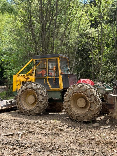 The image shows a yellow and green LKT 81 Turbo forestry tractor parked on a dirt surface near a forest. The tractor's wheels are fitted with heavy-duty chains. The location is likely near the town of Рилски манастир, Bulgaria, given the context.