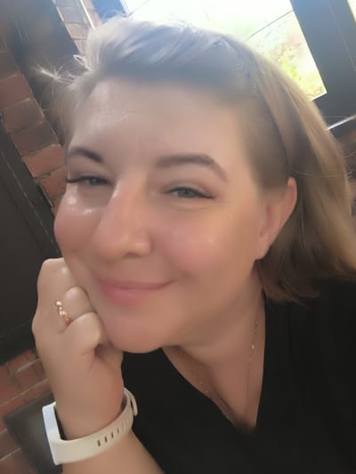 A woman with blonde hair is pictured in a close-up selfie. She is wearing a black shirt and a white watch. Her right hand is resting on her chin and she is wearing a ring on her right index finger. She is smiling and looking towards the camera. The background shows a brick wall and a window with greenery visible outside. The location is not specified in the image.