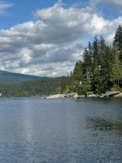 The image shows a body of water with a tree-covered hillside on the right. The hillside is dotted with houses and a tall radio tower is visible in the distance. Several docks extend from the shore into the water. Two people are standing on paddleboards in the water on the left side of the image. The sky is blue with scattered white clouds. This scene is likely in the District of North Vancouver, Canada, given the characteristic landscape of mountains, forests, and waterfront properties.