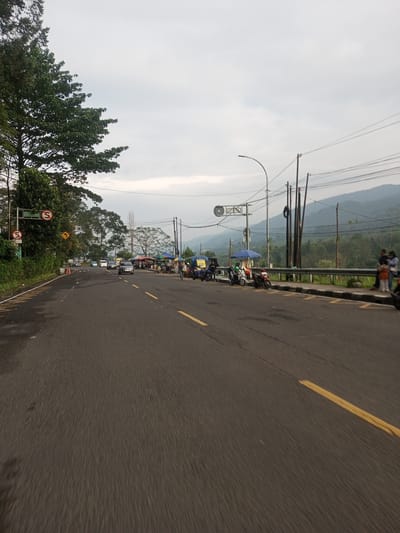 The image depicts a road in a mountainous region. The road is asphalt, with yellow lines marking the lanes. There are trees and greenery on the left side of the road. On the right side, there is a row of vendors and parked motorcycles along the shoulder. In the distance, there are mountains. The sky is overcast. There are some people visible on the right side of the road, possibly interacting with the vendors. The scene appears to be a roadside market area in a rural or semi-rural setting. The location is not Taunggyi, Myanmar.