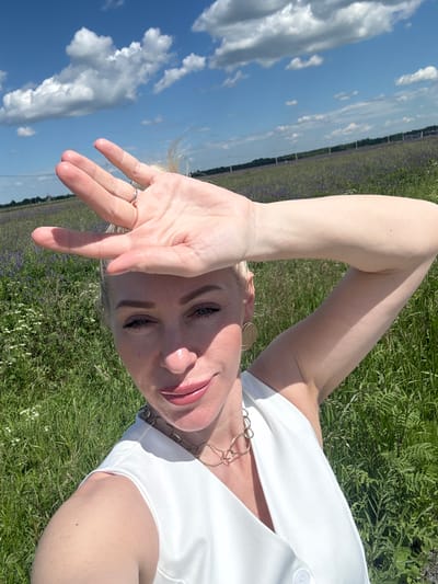 A woman with blonde hair is taking a selfie in a field of purple flowers under a blue sky with white clouds. She is wearing a white sleeveless top and a gold necklace. Her right arm is raised, and her hand is shielding her eyes from the sun. The field extends into the background, with a line of trees and a fence visible in the distance. The scene appears to be outdoors, possibly in the city of Mar'ino, Russia, given the information provided about the location. The lighting suggests it is a sunny day.
