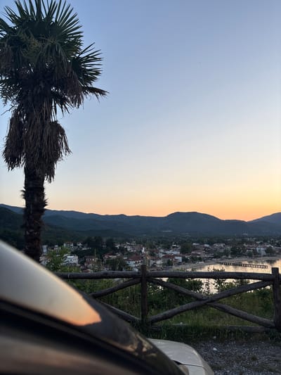 An outdoor shot, rotated 90 degrees counterclockwise, captures a view of Olympiada, Greece, at dusk. A palm tree stands on the left side. The sky transitions from blue to orange behind mountains that slope down to the town. Buildings with red-tiled roofs are visible. A wooden fence runs along the right side of the frame, with a sliver of a vehicle’s body also appearing on the right.