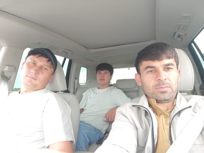 Three men are in the interior of a car, likely an SUV, traveling on a road. The man in the foreground on the right is wearing a tan collared shirt and a light-colored jacket, with his seatbelt visible. The man in the middle is younger, wearing a light-colored t-shirt and blue jeans, and is seated behind the driver. The man on the left is wearing a black baseball cap and a white t-shirt, and appears to be the driver or front passenger. The car's interior, including the ceiling lights, sunroof, and leather seats, are visible. The environment outside the car through the windows suggests a daytime scene with some greenery. The image appears to be a selfie taken by one of the passengers.