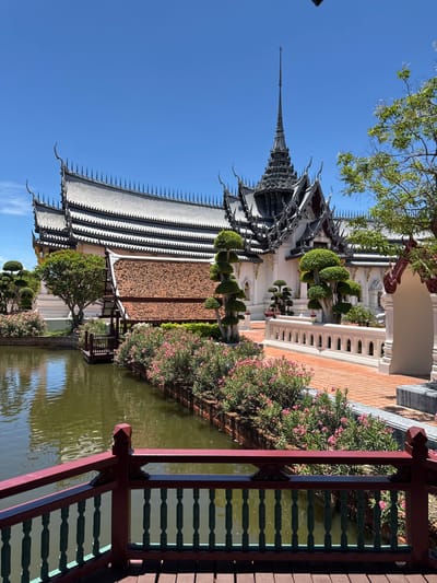 The image depicts Sanphet Prasat Palace, located in the Ancient City (Muang Boran) of Bang Pu Subdistrict Municipality, Thailand. The palace is characterized by its traditional Thai architectural style, featuring multi-tiered roofs with ornate detailing and a central spire reaching towards a clear blue sky. The palace is set against a backdrop of manicured landscaping, including decorative trees, flowering bushes with pink blossoms, and a reflecting pond. A portion of a wooden bridge or walkway with a decorative railing is visible in the foreground.