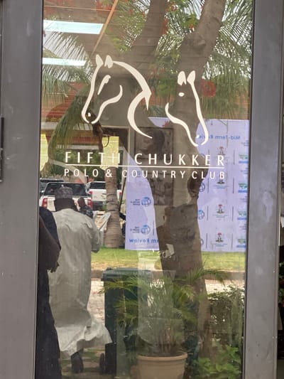 The image is a photo taken through a glass door or window, displaying prominent reflections of an outdoor scene and people, overlaid with branding. A white, stylized logo of two mirrored horse heads, one facing left and one right, is applied to the glass. Below it, the vertical text "FIFTH CHUKKER POLO & COUNTRY CLUB" is also visible in white.
The reflections indicate a bright, sunny daytime environment. Lush greenery, including palm trees and a tree with reddish-orange flowers, is prominent. In the background, a large white banner with repeated logos, such as a red cap/hat icon and a globe icon, and mirrored text (partially legible as "M-BIM" and "weive"), suggests an ongoing event or sponsorship. A grassy area, likely a polo field, and several parked vehicles are also reflected.
Multiple people are visible in the reflections. A person in a light-colored, possibly traditional, garment stands in the lower-middle, with another individual wearing a dark, patterned cap behind them. The scene depicts activity outside the entrance of the Fifth Chukker Polo & Country Club, located in Kwarau, Nigeria.