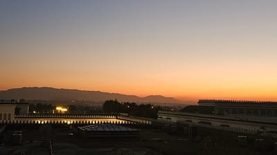 A serene landscape unfolds beneath a gradient sky, transitioning from pale blue to vibrant orange at the horizon. Distant mountains are silhouetted against the glowing sky, while a sprawling cityscape twinkles with scattered lights below. Intricate architectural details, including decorative rooftops and ornate edges, define the foreground, with a flat rooftop area filled with equipment, creating a distinct contrast to the natural beauty beyond.