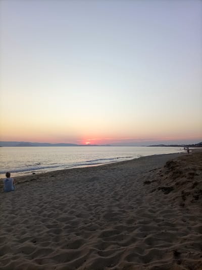 An outdoor, wide-angle shot captures a serene beach scene in Naxos, Greece, at dusk. The sky transitions from a pale blue at the top to soft hues of yellow and pink near the horizon, where the sun is setting behind distant hills. The calm sea reflects the sky's colors, with gentle waves lapping at the sandy shore. In the foreground, a person sits alone on the sand, facing the sea. The beach itself is textured with ripples from the wind. Further down the coastline, a few other individuals can be seen walking or standing by the water. The overall atmosphere is peaceful and tranquil, typical of an evening on a Greek island.