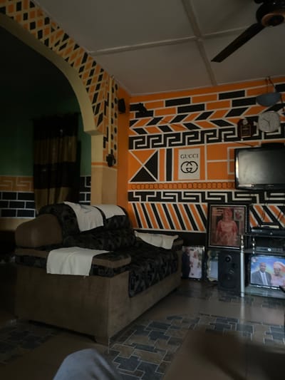 The image displays an interior living space in Uffah, Nigeria, characterized by walls decorated with geometric patterns in orange and black. The prominent right wall features stacked black rectangular blocks, angular black and orange striped sections, and a black Greek key design, with a white stenciled "GUCCI" logo centrally placed. An arched entryway, also adorned with black and orange diamond patterns, connects to an adjacent room where the wall is painted green with a black and orange pattern along its base.
In the foreground, a large, brown upholstered sofa with dark patterned cushions is visible, featuring three white towels draped over its back and seat. Above the sofa, to the right of the archway, a black speaker is mounted on the wall. The ceiling in the upper right reveals a dark ceiling fan with two blades and a white circular analog clock mounted on the wall below it.
A television set rests on a black multi-tiered shelf unit to the right. Below and around the television, several framed photographs are displayed, including one of a woman in traditional attire and another showing a wedding couple. A black audio speaker stands on the floor to the left of the television stand. The floor is covered with patterned tiles in shades of brown and gray. The room is dimly lit, and no individuals are actively present in the scene.