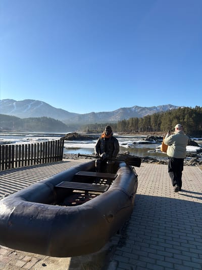 The image depicts a sunny outdoor scene in Manzherok, Russia, featuring a statue of a river raftsman on a metal contraption that resembles a raft, positioned along a brick walkway. In the background, a mountain range rises under a clear blue sky. A body of water, possibly a river, is visible with ice floes. A person wearing a light-colored jacket and dark pants is standing on the walkway, appearing to take a photograph. The scene is captured in a landscape orientation.