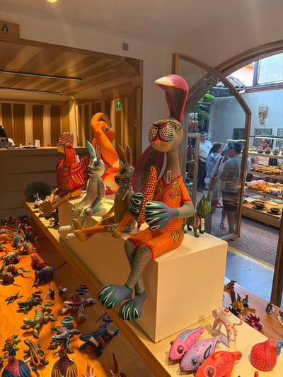 The image showcases a collection of colorful, intricately designed alebrijes, folk art sculptures, arranged on a wooden shelf. Dominating the foreground is a rabbit-shaped alebrije with vibrant orange and green accents. Smaller alebrijes, including foxes and birds, are stacked beneath it. A mirror in the background reflects the store's interior, revealing a shopper. Other alebrijes, in the shapes of fish and other animals, line the shelf, creating a whimsical display.