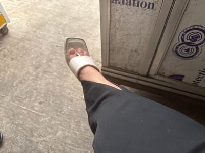 In the city of Meunasahmanyet, Indonesia, a person's left foot is visible, clad in a white slip-on sandal with a square toe. The foot is resting on a textured, light-colored surface, possibly concrete or linoleum. Dark, wide-leg pants are worn, with a slit detail visible near the ankle. In the background, a metallic structure with some text and a circular logo is partially visible, suggesting an urban or commercial setting. The lighting appears to be natural, casting subtle shadows.