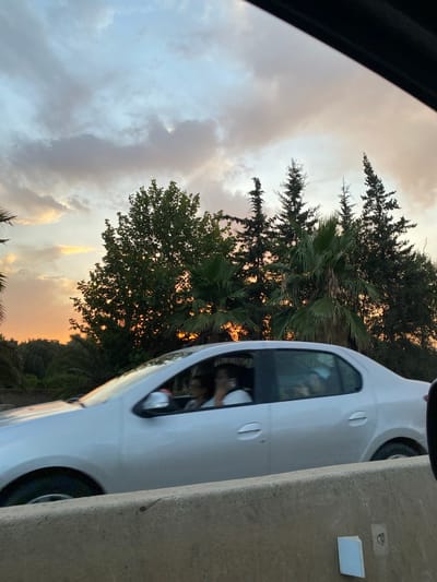A silver sedan is captured in motion, its occupants blurred due to the speed and lighting. The car is traveling on a road, with a concrete barrier visible in the foreground. Behind the car, a dense collection of trees, including palm trees and what appear to be cypress trees, forms a lush backdrop. The sky above is a mix of soft clouds, transitioning from a pale blue to warm hues of orange and yellow, indicating either sunrise or sunset. The overall impression is a snapshot of a journey through a scenic, possibly rural, area near El Karia, Algeria, characterized by its natural vegetation and the soft glow of twilight.