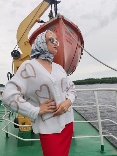 A woman stands on the deck of a boat, looking to her right. She has short, pink hair and is wearing white sunglasses, a patterned silk scarf tied around her head, and a white cardigan with light brown heart patterns. Beneath the cardigan, a red skirt is visible. Her left hand is on her hip, and her right hand is by her side. Behind her is a red lifeboat, suspended from a yellow crane. The boat is on a body of water, with a tree-lined shore in the background. The sky is overcast, suggesting a cloudy day. The visible text on the lifeboat reads "NAKHODKA" and "CANTON". The scene appears to be in Bereg, Russia.