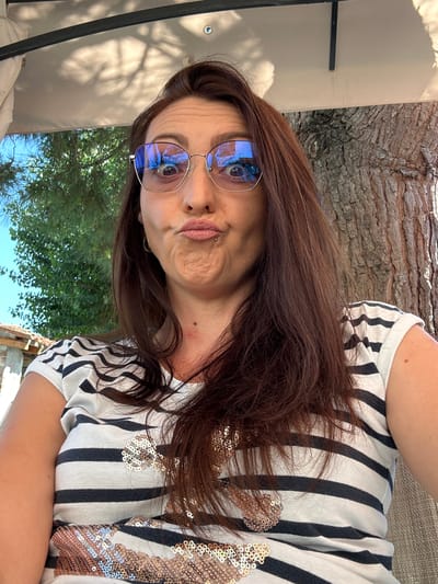 A woman with long, brown hair and blue-tinted glasses makes a duck face while taking a selfie outdoors. She is wearing a striped t-shirt with sequined embellishments on the front. Behind her, a large tree trunk is visible to the right, and greenery and a clear blue sky are to the left. The scene appears to be in a garden or park setting.