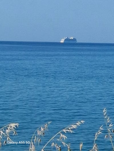 The image shows a view of the sea or a large body of water, likely taken near the Gouves Municipal Unit in Greece based on the context provided. A portion of the sky is visible on the left side of the image, along with some foliage or tall grass in the right foreground. The sea appears calm, indicated by the texture of the water in the image. The colors are dominated by shades of blue and white. The text "Galaxy A55 5G" is visible at the bottom right corner.
