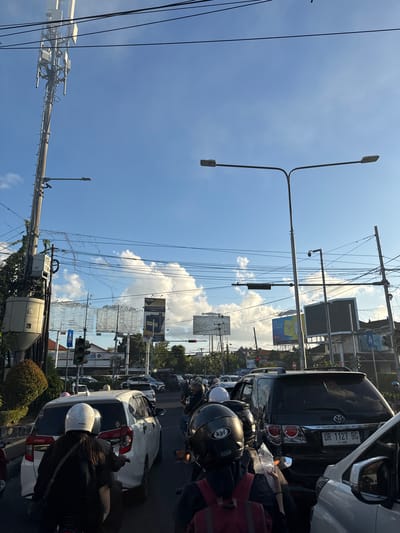 The image depicts a traffic jam on a sunny day, likely in the city of Denpasar Barat, Indonesia. The scene is dominated by various vehicles including cars and motorcycles, with many individuals wearing helmets. The sky is blue with scattered white clouds. Prominently visible are utility poles with numerous wires, a cell tower, and streetlights. Several billboards are present in the background, some displaying advertisements, including one for "NEW MODERN KRETEK". Traffic lights are also visible. The overall impression is of a bustling urban environment during daylight hours.
