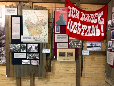 This image shows a museum exhibit in Ust'-Pyzas, Russia, featuring historical artifacts and documents related to the Russian Revolution and Civil War. The exhibit is displayed on a wooden wall, with various items mounted at different heights. In the upper left, a white sign with black text reads "Ц. ... ПО ТРАКТУ МНОГО ИДЕТЬ БРОДЯТЬ И ПРОЛЕТАРНЕВЪ БОСЬКЪ И ПОЛЬЗК ИЗЪ ССЫЛКМЫ - НЕСОГЛАСИЕМЪ И ДАКО ИЗГОТОВЛЕНИЙ." Below this is a black board with three documents. The top document is titled "УСТАВЪ," followed by smaller text. The second document is also titled "УСТАВЪ" with more text. The third document is smaller and appears to be a typed letter. To the right of these documents is a large, faded map titled "КАРТА ТОМСКОЙ ГУБЕРНИИ." The map depicts geographical features and colored regions, likely representing administrative divisions or resources. Below the map is a sign with text in black. Dominating the upper right portion of the image is a large red banner with white Cyrillic lettering. The text appears to be a revolutionary slogan. Below the banner, a dark rectangular plaque displays a bust of a man and a smaller portrait of a person in uniform. A long, wooden rifle, possibly a Mosin-Nagant, leans against the wall vertically in the center of the exhibit, its green strap visible. To the right of the rifle, a dark curved sword is mounted diagonally. Below the rifle and to the left, a black board contains several black and white photographs arranged in a grid. The top row has three images, and the bottom row has one larger image. Below these photos are two metallic objects: a set of handcuffs and a key with a chain. To the right of the rifle and sword, there is another display. At the top is a black and white photograph of a group of people standing in front of a building. Below this are two more photographs. One shows a group of people in a snowy landscape, and the other shows a single person in uniform. Further to the right, a red board has a title in large Cyrillic letters: "КАК И ПОЧЕМУ ПОБЕДИЛА СОВЕТСКАЯ СТРАНА ИНТЕРВЕНЦИЮ И КОНТРРЕВОЛЮЦИЮ В РОССИИ." Below this title are two more black and white images depicting scenes of conflict or gathering. Several smaller white signs with black text are placed throughout the exhibit. One sign, positioned between the rifle and the red board,