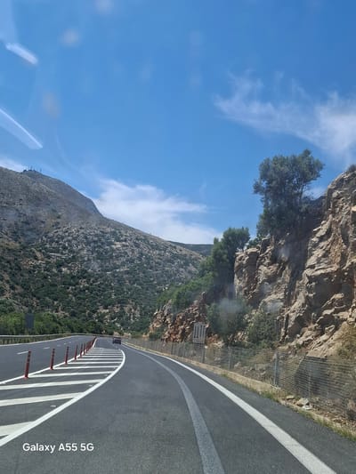 The image depicts a view from inside a vehicle traveling along a winding road. The road is characterized by white lane markings and vertical red and white posts along the shoulder. To the right of the road, there is a steep, rocky mountain covered in vegetation. A wire mesh is visible along the edge of the road, likely for rockfall protection. The sky is blue with scattered white clouds. The photograph was taken with a Samsung Galaxy A55 5G, as indicated by the watermark. The location is likely within the Gazi Municipal Unit, Greece, given the typical mountainous landscape and road construction. The photo does not depict any people.