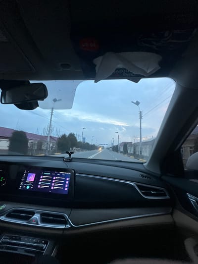 The image depicts a street view from inside a moving or stationary vehicle in Yangiabad, Uzbekistan, during what appears to be dusk or dawn. The front windshield frames a two-lane asphalt road stretching into the distance, lined with streetlights featuring rectangular LED fixtures on utility poles. On the left side of the road, residential buildings with reddish-brown roofs are visible, interspersed with trees. On the right, lighter-colored buildings or walls are present, also with trees and utility lines overhead. A few distant vehicles with illuminated headlights are visible on the road.
Inside the vehicle, the dark-colored dashboard occupies the lower half of the frame. A large infotainment screen centrally displays a music application interface with album art and song titles in purple and blue tones. Below this, air vents and climate controls are visible. A small black device, possibly a phone mount or dashcam, is attached to the dashboard near the base of the windshield. The rearview mirror is visible at the top-center of the windshield. Above the windshield, a red circular object with "70 07" text and a crumpled white tissue are attached to the overhead lining, with a partial "Qolla Billa" label also visible. No individuals are explicitly visible.