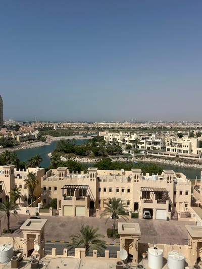 The image captures an aerial view of a developed area with buildings, a waterway, and lush greenery. The buildings are mostly modern, with several floors and beige exteriors, featuring balconies, windows, and air conditioning units. Some buildings are clustered together, forming a residential or commercial complex, while others are more spread out. A prominent feature is the waterway that winds through the landscape, bordered by stone walls and vegetation. Palm trees are scattered throughout the scene, adding a tropical feel. In the foreground, several large white water tanks are visible on rooftops, along with satellite dishes and air conditioning units. A white car is parked in an open garage area. The overall setting appears to be a well-maintained, affluent area, possibly a resort or residential community. The sky is clear and blue, suggesting a sunny day. The image is oriented vertically, as if taken from a high vantage point looking down. The location is identified as Al Jazeer Heritage Village in the United Arab Emirates. There is no discernible text in the image.