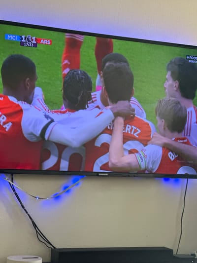 The image shows a television screen displaying a football (soccer) match between Manchester City (MCI) and Arsenal (ARS), with the score tied at 1-1 and the game clock at 17:33. The television is a Thomson brand. On the screen, Arsenal players are celebrating, as they are depicted embracing each other. The jerseys are red and white, and one player has a captain armband. The wall behind the TV is off-white. There are black cables and a roll of toilet paper adjacent to the TV. Blue LED lights are attached behind the television. The location of the image is unknown, but the user indicated a reference to the city of Nogent-sur-Oise, France.