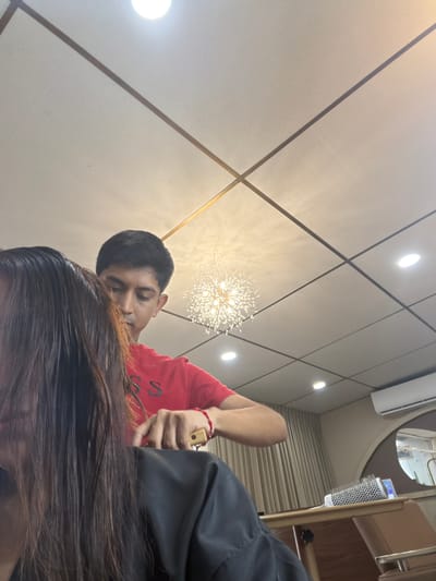 A medium shot depicts a man with dark hair, wearing a red t-shirt and a red bracelet, cutting long brown hair. The setting appears to be indoors, possibly a hair salon, under bright artificial lighting. The walls are beige with thin brown lines forming squares, and a decorative light fixture is visible. The scene suggests a hair styling session in progress. Location is in Villahermosa, Mexico.