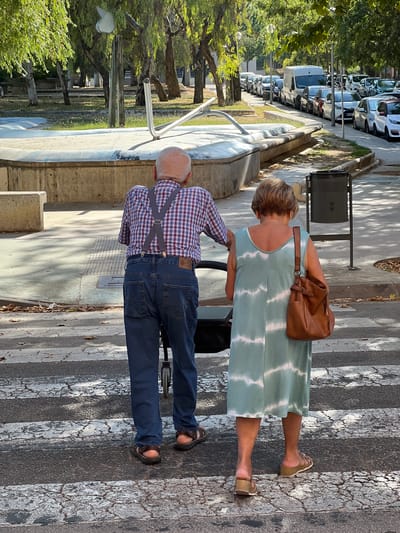 Two people, an older man and woman, stand on a crosswalk in Mollet del Vallès, Spain. The man, wearing a red and blue plaid shirt, denim jeans, and grey suspenders, is facing away from the camera. The woman, in a light blue and white tie-dye dress and brown sandals, is also facing away and has a brown leather purse slung over her shoulder. Her hair is short and blonde. The sun is casting shadows on the ground, indicating it is daytime. The scene appears to be a street with trees lining one side and parked cars in the background. The crosswalk stripes are worn.