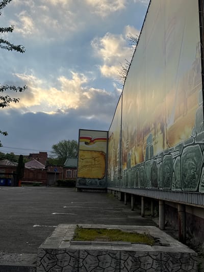 The image shows a parking lot in Akhieli, Georgia, adjacent to a large billboard and a stone wall. The billboard is elevated, with a design resembling a historical panorama and a smaller sign beneath depicting a map or historical document. Buildings, including a brick structure, are visible behind the billboard. The sky is partially cloudy. The parking lot is marked with white lines, and a section of the stone wall is topped with green vegetation.
