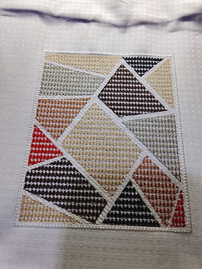 The image shows a close-up of a textile with an embroidered geometric pattern. The pattern is square and composed of various triangles filled with different colored threads, including shades of brown, beige, red, black, and gray. The embroidery creates a textured effect with small, repeating stitches. The textile itself is white and appears to be a coarse weave, possibly a linen or cotton blend. The embroidered section is bordered by a white stitched edge, suggesting it might be an appliqué or a patch sewn onto the larger fabric. The background fabric is slightly rumpled, indicating it's a flexible material. There is no visible text in the image, and the lighting suggests it was taken indoors or in diffused natural light. The context of Rumuepirikom, Nigeria, does not provide specific visual cues within the image itself.