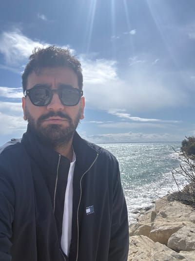 A single male individual is depicted in a daytime outdoor setting in Agios Tychonos, Cyprus. He is positioned in the foreground, facing directly towards the camera, from the chest up. The man has dark hair and a full beard, and is wearing dark-framed sunglasses, a dark blue or black zip-up jacket with a "TOMMY JEANS" logo on the left chest, over a white t-shirt. The background features a body of water, consistent with the Mediterranean Sea, exhibiting sunlight reflections on its surface and a rocky shoreline on the right. Sparse, dry vegetation is visible on the rocks. Above the horizon, a partly cloudy blue sky is present, with the sun casting prominent rays from the upper left quadrant. A distant structure or landform is faintly visible across the water on the horizon.