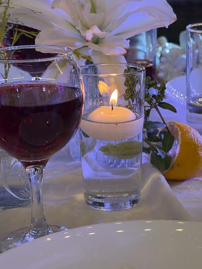 The image depicts a close-up view of a table setting. In the foreground, a stemmed wine glass filled with red wine is prominently displayed on the left side. To its right, a tall cylindrical glass holds water with a floating white candle illuminated, casting a warm glow. A sprig of greenery with small green berries is placed next to the candle. A halved yellow lemon with some leaves attached rests on the right side of the frame. In the background, a large white flower with multiple petals dominates the scene, and out-of-focus elements suggest other glassware and decorative items. The lighting is dim and atmospheric, with blue and white tones prevalent, creating a soft, elegant ambiance. The setting appears to be indoors, possibly for a special occasion or meal. The location in the city of Bereke, Kazakhstan, is not discernible from the visual information presented in the image.