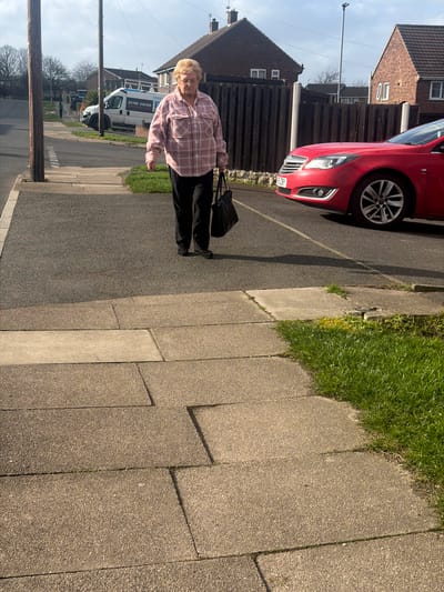 The image displays a daytime residential street scene located in the city of Rotherham, United Kingdom. An elderly Caucasian female, with short blonde hair and glasses, walks towards the camera on a paved concrete sidewalk. She is dressed in a pink and white plaid long-sleeved shirt, black trousers, and dark flat shoes, carrying a black bag in her left hand.
The sidewalk, constructed from large light-colored concrete slabs, abuts a dark asphalt road. A red car, with a partial UK license plate "56 ZBY" visible on its front, is parked on the left side of the road, partially on a grass verge, with its front section visible. Further down the road on the left, a white panel van, bearing blue lettering including "DOOR J..." and the phone number "01709 516100", is parked. A second, less distinct individual is visible walking in the distance on the left-side pavement.
In the background, several brick residential houses are present, separated from the road by fences, one of which is a dark wooden fence with concrete posts. Two dark utility poles are positioned on the left side of the street. A modern street light pole is also visible in the mid-ground. The environment includes green grass verges and bare trees in the distance. The sky is partially cloudy, and the scene is illuminated by natural daylight, casting shadows.