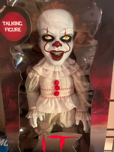 The image shows a Pennywise the Dancing Clown talking figure in its original packaging. The figure is dressed in a white costume with red trim and three red pom-poms down the front. It has a white face with red makeup, yellow eyes, and a wide, grinning mouth. The packaging is clear plastic with a dark cardboard backing that features an image of trees. A red oval sticker on the left side of the packaging reads "TALKING FIGURE" in white text. The figure is positioned centrally within the frame. No specific location in Schwäbisch Hall, Germany can be determined from the image.