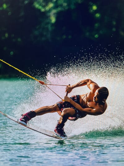 A shirtless man is wakeboarding on a body of water, likely a lake, with trees visible on the far left. The man is wearing patterned shorts and boots attached to the wakeboard. He is holding onto a wakeboarding rope and handle. Water droplets are spraying up from the wakeboard and the surrounding water, suggesting movement and speed. The location is Bacong, Philippines.