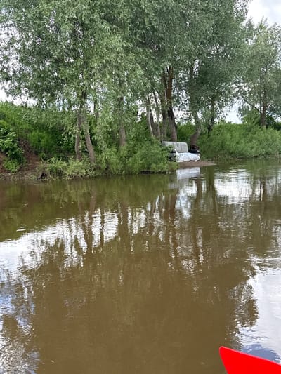 The image shows a river scene, possibly the location of Izh-Boyki, Russia, with trees and vegetation lining the riverbank. A red object is visible in the water, and there appears to be debris or objects (possibly bags) near the shoreline. The water surface is reflective, showing the sky and vegetation.