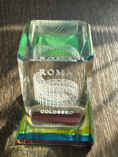 This is a close-up, overhead shot of a crystal souvenir with colored segments on the right side. The souvenir is resting on a wooden surface that is dappled with sunlight and shadow, suggesting an outdoor setting during the day. The crystal appears to be shaped like a rectangular prism with faceted edges. Inside the crystal, there are etched images. On the left, there's a circular emblem. In the center, an image of the Colosseum is visible, with the text "ROMA" above it and "COLOSSEO" to its right. The souvenir is predominantly clear with green and blue accents. The light creates a lens effect, magnifying the textured wood grain of the surface. The overall impression is one of a captured memory, a small piece of Roman heritage.