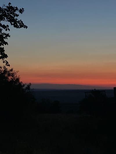 The image shows a landscape at dusk. The sky transitions from a deep blue at the top to shades of orange and red near the horizon. Silhouetted trees and foliage frame the scene from the left and right, with a dark, indistinct landscape visible in the middle ground. The location is specified as Cairaclia, Moldova. The image is a still photograph, depicting a natural environment with no human subjects or discernible activity. The overall impression is one of a peaceful, natural setting at twilight.