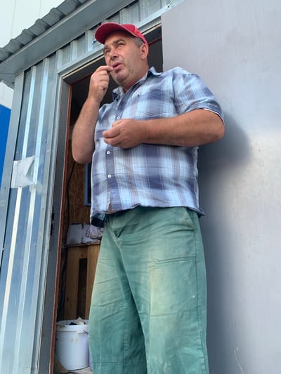 A middle-aged man with fair skin and a bald head, wearing a red baseball cap, a blue and white plaid collared shirt, and faded green work pants, stands in the doorway of a corrugated metal structure. He is looking up and to his left, with his right index finger and thumb near his mouth, as if picking at something. His left hand is loosely clenched. The structure appears to be a utilitarian outdoor building or shed, with a light-colored wall on the right. Inside the doorway, visible items include a white bucket and some wooden shelving. The lighting suggests it is daytime, with shadows cast on the wall. This scene is located in Pivdennoukrainsk, Ukraine.