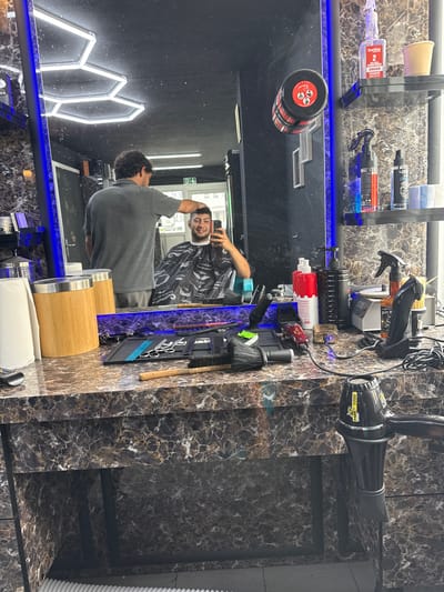 This image depicts the interior of a modern barbershop in Lausen, Switzerland, centered around a barber station. The main subject is a man, seen from behind in a grey t-shirt, with dark curly hair. He is looking into a large mirror, which reflects his smiling face. In the reflection, he is wearing a black barber's cape adorned with white mustache patterns, suggesting he is either preparing for or has just received a haircut.
The barbershop features a counter and back wall with a distinctive dark, marbled pattern. A blue LED strip provides ambient lighting along the counter's edge and under a floating shelf above it. On this shelf, several hair products are visible, including a "borthe 2 PHASE CONDITIONER ARGAN" bottle containing purple liquid, a black bottle partially labeled "CODE," and a yellow paper cup.
The counter below is well-stocked with a variety of barber tools and supplies. These include hair clippers, spray bottles (one clear with "Just W" and brown liquid, another black), brushes, combs, and a black hair dryer labeled "PARS 5000" and "1000W." A red and white "Color Up Turbo" clipper is also present, along with a black cylindrical container with a red lid, possibly holding hair styling product. Charging cables and adapters are visible, powering some of the electronic devices. On the right, two wooden cylindrical containers with silver tops and a roll of white paper towels complete the setup. A black rubber mat neatly organizes several tools, including scissors.
In the background reflected in the mirror, white hexagonal LED lights are mounted on a dark wall, contributing to the contemporary aesthetic of the space. The man's smiling reflection suggests a positive and relaxed atmosphere.