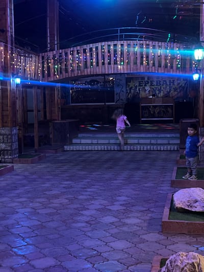An outdoor courtyard area features a child in a blue t-shirt and grey pants standing on a raised platform with grass. Another child in a pink t-shirt and light-colored shorts is running across a stage. The stage area is lit with colorful lights. Behind the stage, a banner displays the word "ЕРЕВАН" in large letters, with silhouettes of people dancing. The courtyard is paved with interlocking stones, and decorative lights are strung along a wooden railing. Large rocks are placed in separate sections of the courtyard. The scene is illuminated by blue and purple lighting.