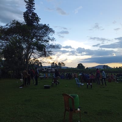 The image shows a gathering of people outdoors, likely in the Keyian ward, Kenya, suggested by the environment. The setting is a grassy field with a large tree on the left side of the frame. A group of people are standing near the tree, while others are seated in chairs spread across the field. The people appear to be of varying ages. Objects such as plastic chairs, a plastic water container on top of a chair, and a few black metal tubs are scattered around the area. In the background, there are more trees and a horizon with visible clouds. The sky is light blue with patches of white and grey clouds. The overall lighting suggests it is either late afternoon or early evening. There are individuals walking or standing among the seated people.