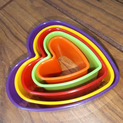 The image displays a nested arrangement of five heart-shaped cookie cutters in graduating sizes, placed on a wooden surface. From largest to smallest, the cutters are purple, yellow, red, light green, and orange. The wooden surface has a planked appearance with visible grain and appears to be a table or countertop. The lighting suggests an indoor setting. There are no people or animals visible in the image. The scene is devoid of any context related to the city of El Limón, Venezuela, or any other specific geographic location.