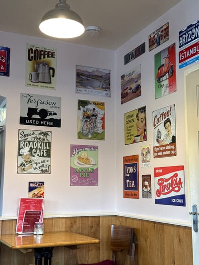The image shows the interior of a cafe or restaurant. The walls are decorated with a variety of vintage-style metal signs featuring advertisements for coffee, tea, and various brands like Pepsi-Cola and Bugatti. The signs also depict tourist destinations like the Lake District and Tour de France. A light fixture is visible on the wall. The setting is the Old Pump House Cafe in Bowness-on-Windermere, United Kingdom. A wooden table is visible, covered with menus and a jar of condiments.