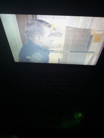 The media file displays a man on a laptop screen, captured from above. The man, who appears to be in his early twenties, has short, dark hair and is wearing a quilted, dark blue jacket with a diamond pattern on the sleeves. A small, green logo with white letters, possibly "HTB", is visible on the left sleeve. He is looking towards the right, with his face partially visible. The background on the screen is blurred and appears to be an indoor setting with shelves or partitions.
Below the laptop screen, the keyboard is visible, mostly in darkness. There are faint green and red lights reflecting on the keyboard area, suggesting the laptop is on and possibly in use. The overall ambiance is dim, indicating the photo was taken in low light conditions. No specific time of day or weather conditions are discernible. The setting seems to be an indoor environment, likely a room where a laptop is being used, with the screen displaying a video or image of the man. No other specific location cues are present besides the provided context of Nova Ivanivka, Ukraine.