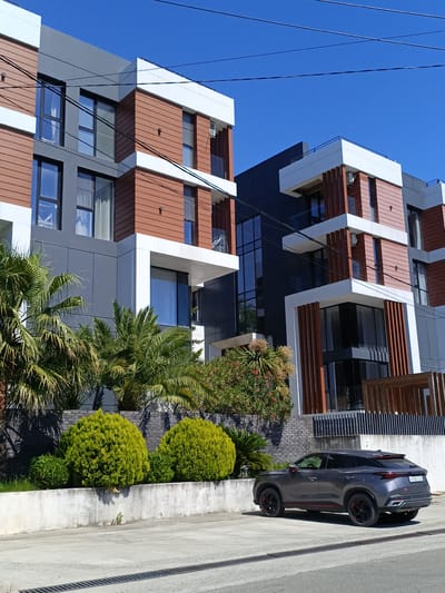 A dark gray crossover SUV is parked on a paved surface in front of a modern apartment building complex in Verkhnevesyoloye, Russia. The building has a distinctive design with alternating sections of gray panels and wooden planks, accented by white framing. Large windows are visible on multiple levels, and balconies overlook the front. Lush palm trees and manicured green shrubs surround the building. The sky is a clear, bright blue. Overhead, several utility wires crisscross the frame. The car's license plate is partially obscured but appears to have Russian characters. The scene appears to be a residential area on a sunny day.