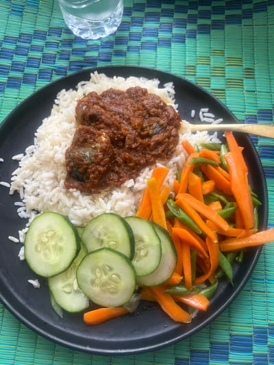 A black plate holds a meal of white rice topped with a dark red stew, alongside a colorful mix of sliced carrots, green beans, and cucumber rounds. A golden spoon sticks vertically out of the rice. The meal rests on a blue and green woven surface. A glass of water is visible in the lower left corner. The food is presented simply and healthily.