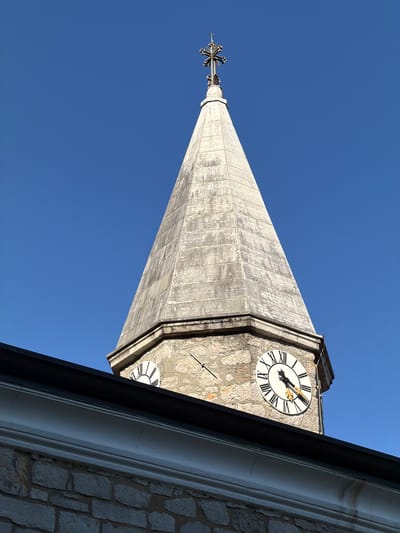 A low-angle shot of the St. James church tower in Grad Opatija, Croatia, highlights its architectural details against a clear blue sky. The tower is constructed from stone, with a clock face visible near the top, displaying Roman numerals. The tower's spire is a light gray, gradually tapering to a point, with a cross-shaped weather vane. A portion of the church's roof and exterior wall is visible in the bottom right corner, showing the stone construction and molding.