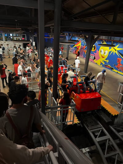 The photograph captures a queue of people waiting for a ride, likely a rollercoaster, at what appears to be a theme park attraction. A partially visible ride vehicle, red with rows of seats, is moving along the track. The surroundings are indicative of an indoor amusement area, with painted walls featuring cartoonish characters and steel support structures. The location is possibly Anaheim, United States of America, based on the theme park-like setting.